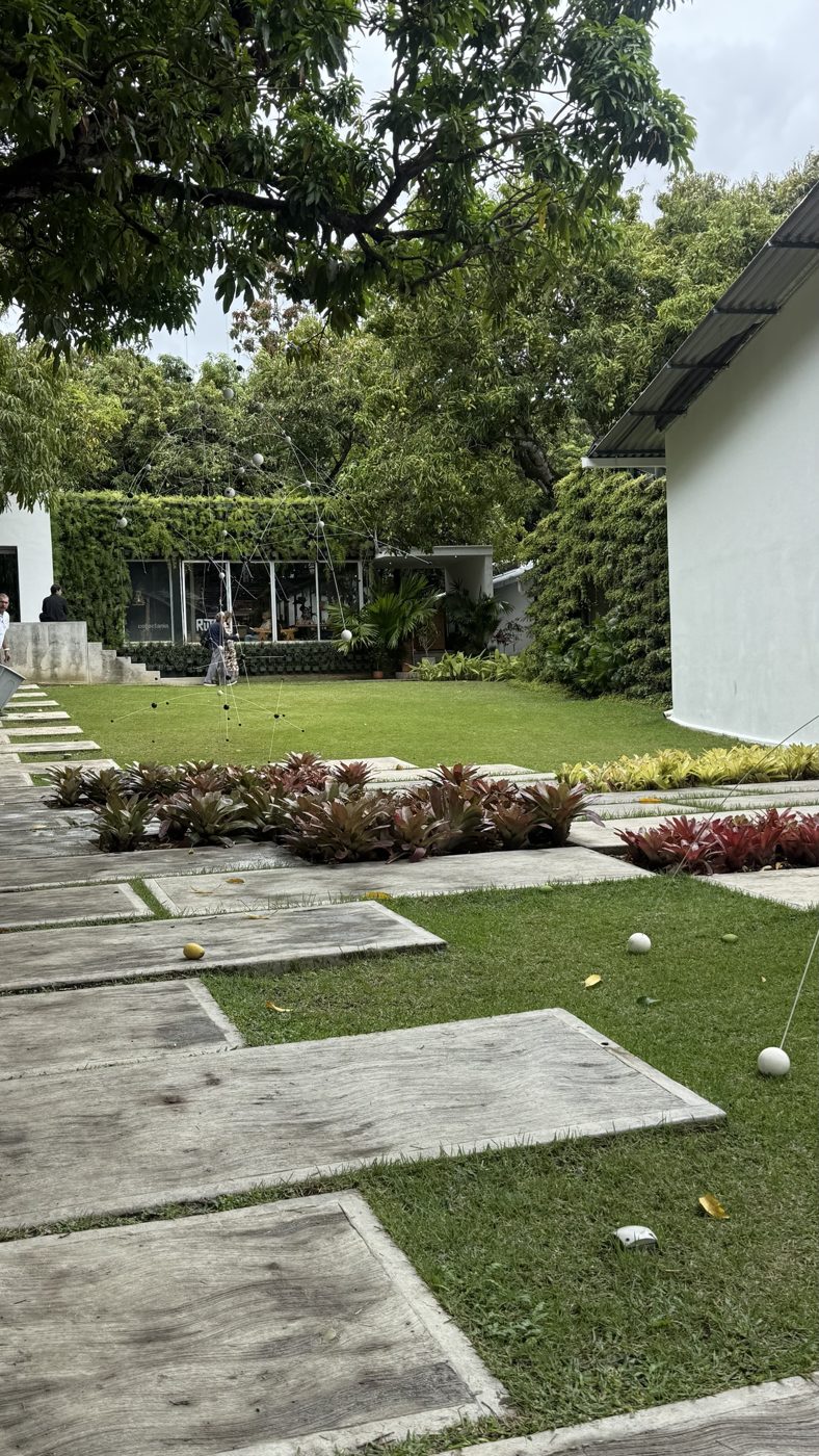 Jardín de Los Galpones con caminos de piedra, bromelias, una escultura de esferas y galerías blancas bajo los árboles